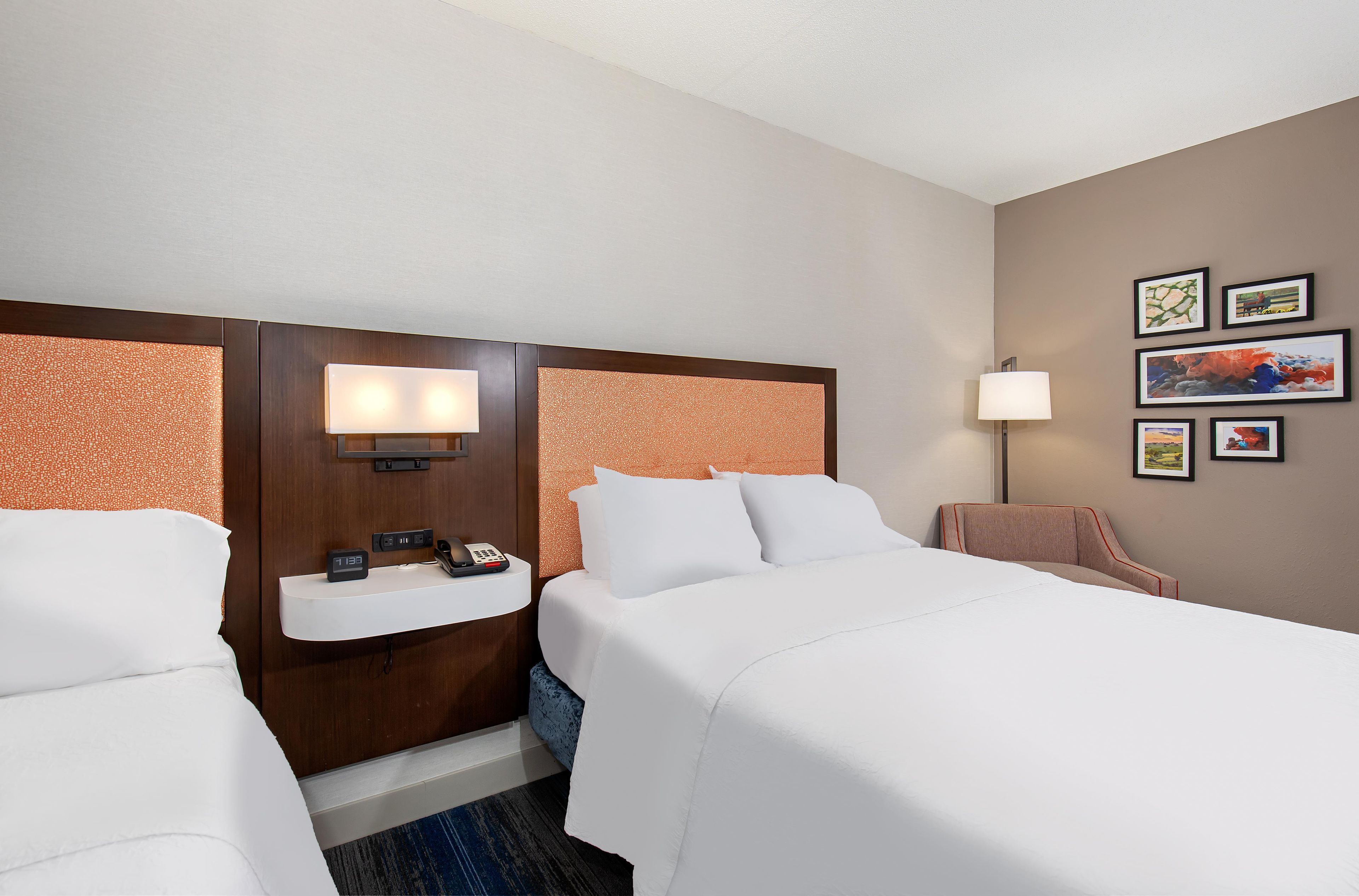 Ein Hotelzimmer mit zwei Betten, einem Nachttisch mit Telefon und Uhr, einer Wandlampe und gerahmten Kunstwerken an der Wand.