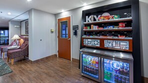 Un hall d’hôtel doté d’un réfrigérateur rempli de boissons, d’une étagère avec des en-cas et d’un coin salon avec une lampe.