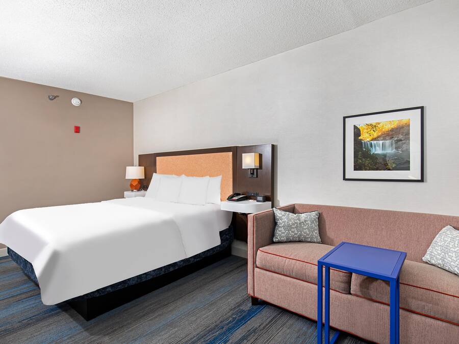 Habitación de hotel con cama, sofá, una mesita, lámpara de pared, un cuadro y termostato.