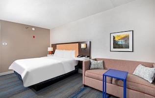 Habitación de hotel con cama, sofá, una mesita, lámpara de pared, un cuadro y termostato.