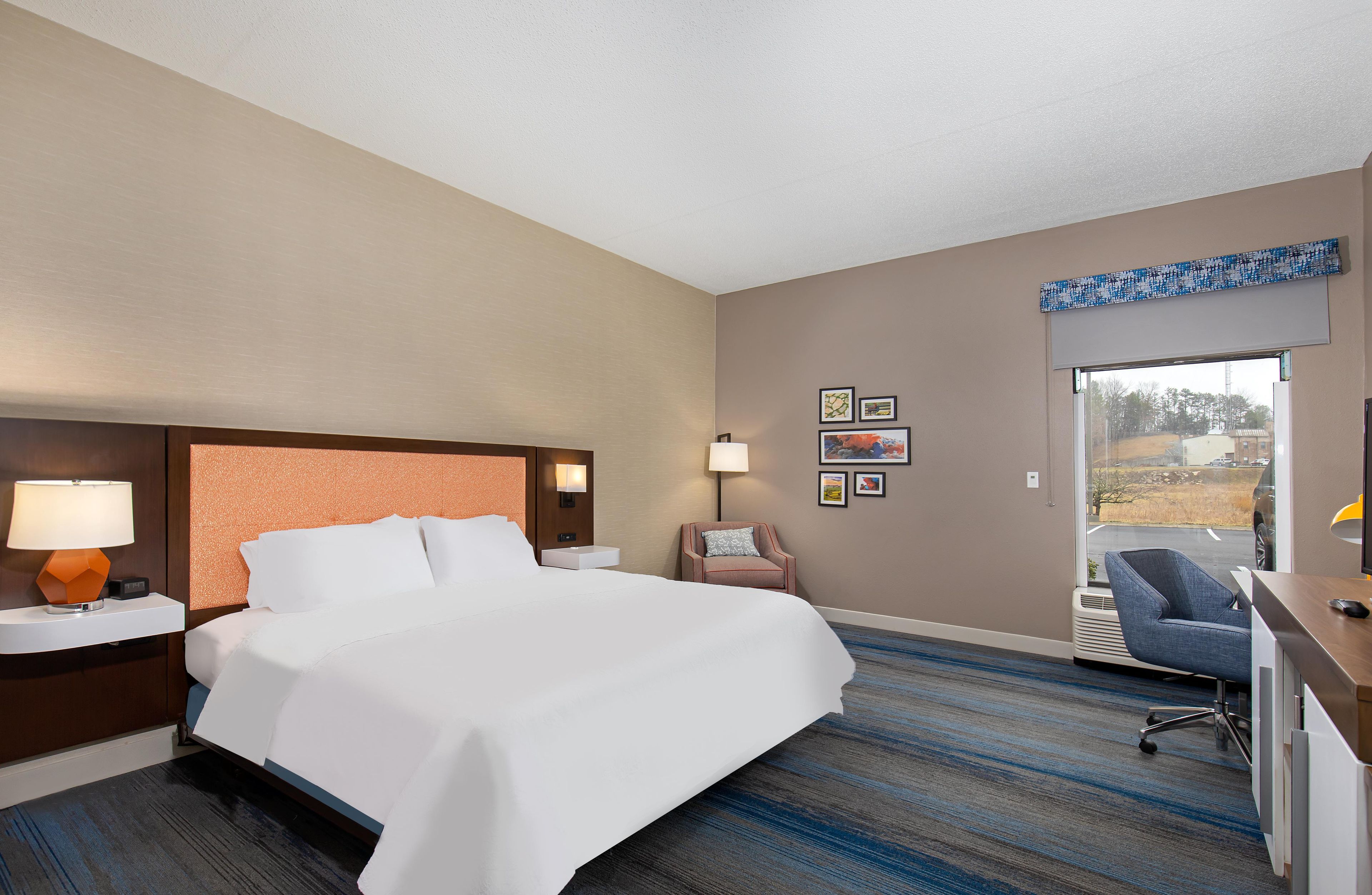 A hotel room with a large bed, two bedside tables with lamps, a chair, a desk with a chair, and a window with a view of the outside.