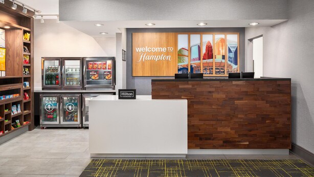 Ein moderner Hotel-Empfangsbereich mit einem Holzbüro, einem 'Welcome to Hampton' Schild und einer Stadtwandmalerei.
