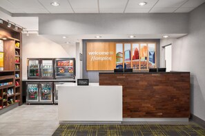 A modern hotel reception area with a wooden reception desk, a 'Welcome to Hampton' sign, and a cityscape mural. - Hampton Inn & Suites Charlotte-Arrowood Rd. (Charlotte)