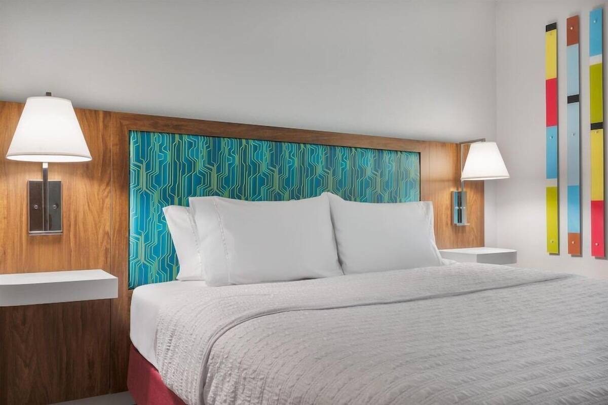 A hotel room with a wooden headboard, white bedding, and a colorful abstract wall art piece.