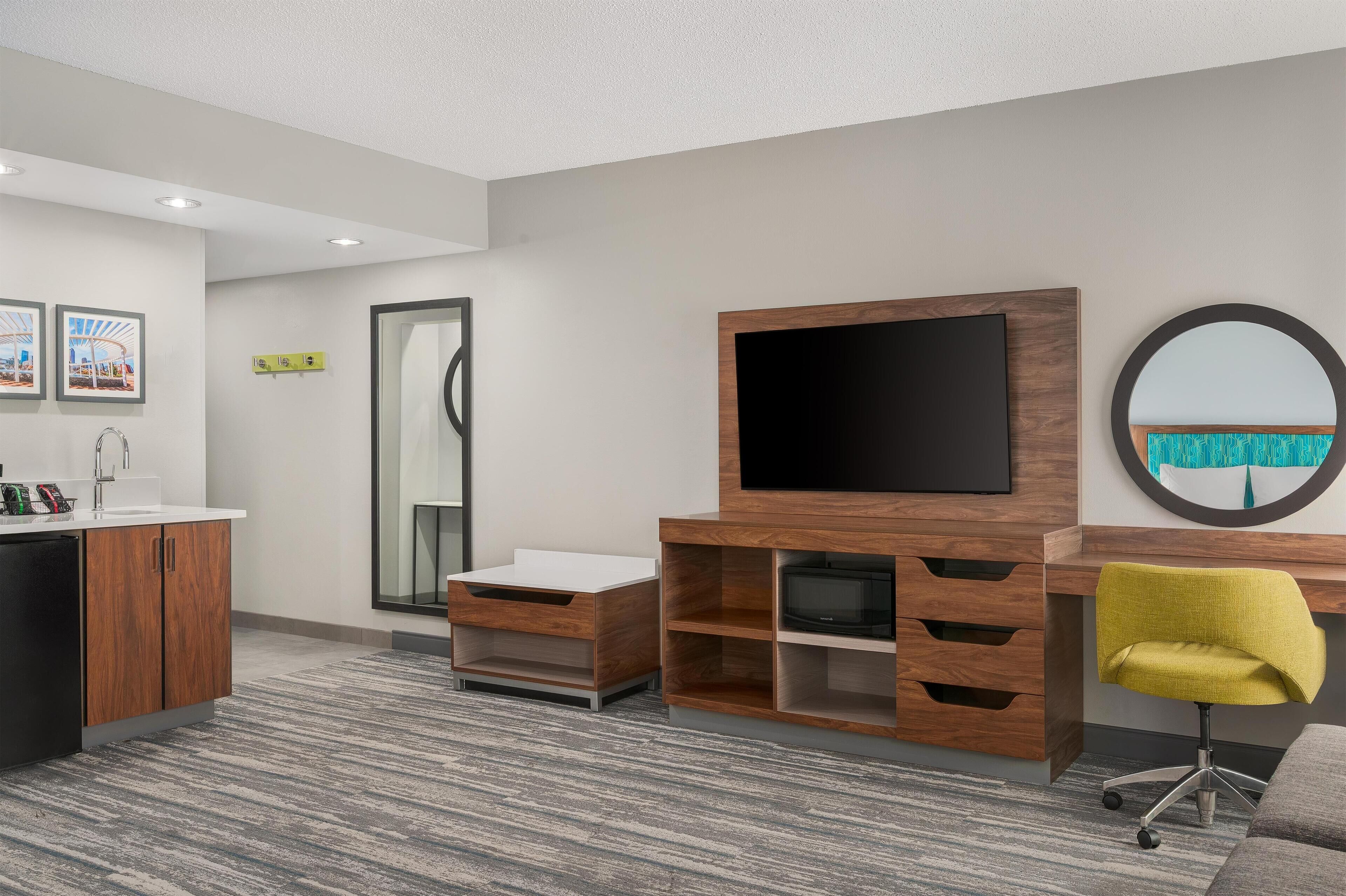 A modern hotel room with a wooden desk, a flat-screen TV, a round mirror, and a yellow chair.
