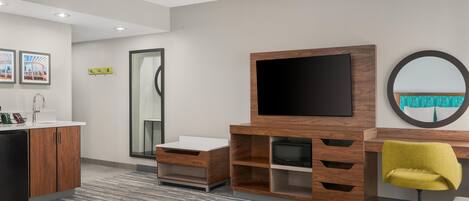 A modern hotel room with a wooden desk, a flat-screen TV, a round mirror, and a yellow chair.