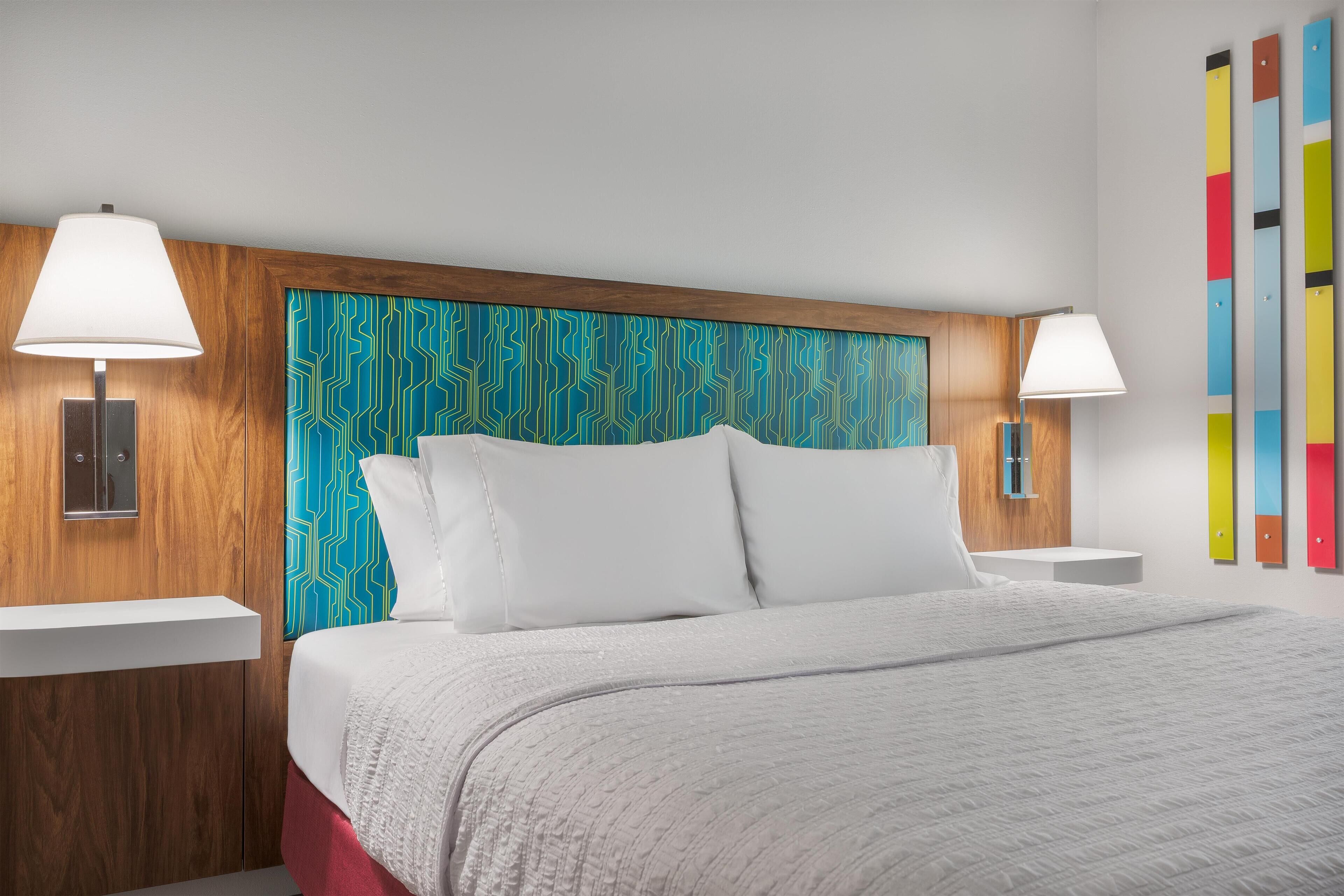 Quarto de hotel moderno com cabeceira de madeira, roupa de cama branca e duas luminárias de cabeceira.