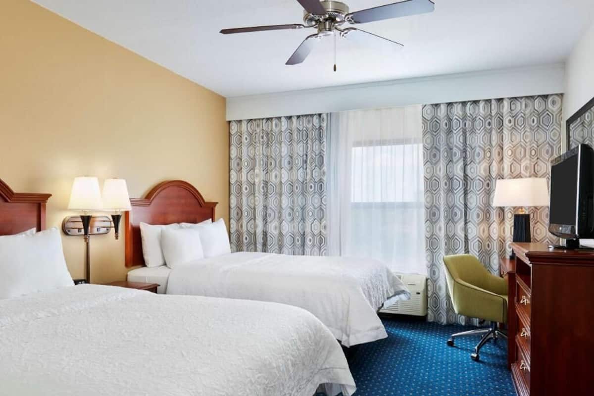 A hotel room with two beds, a ceiling fan, a television, a desk, and a chair.