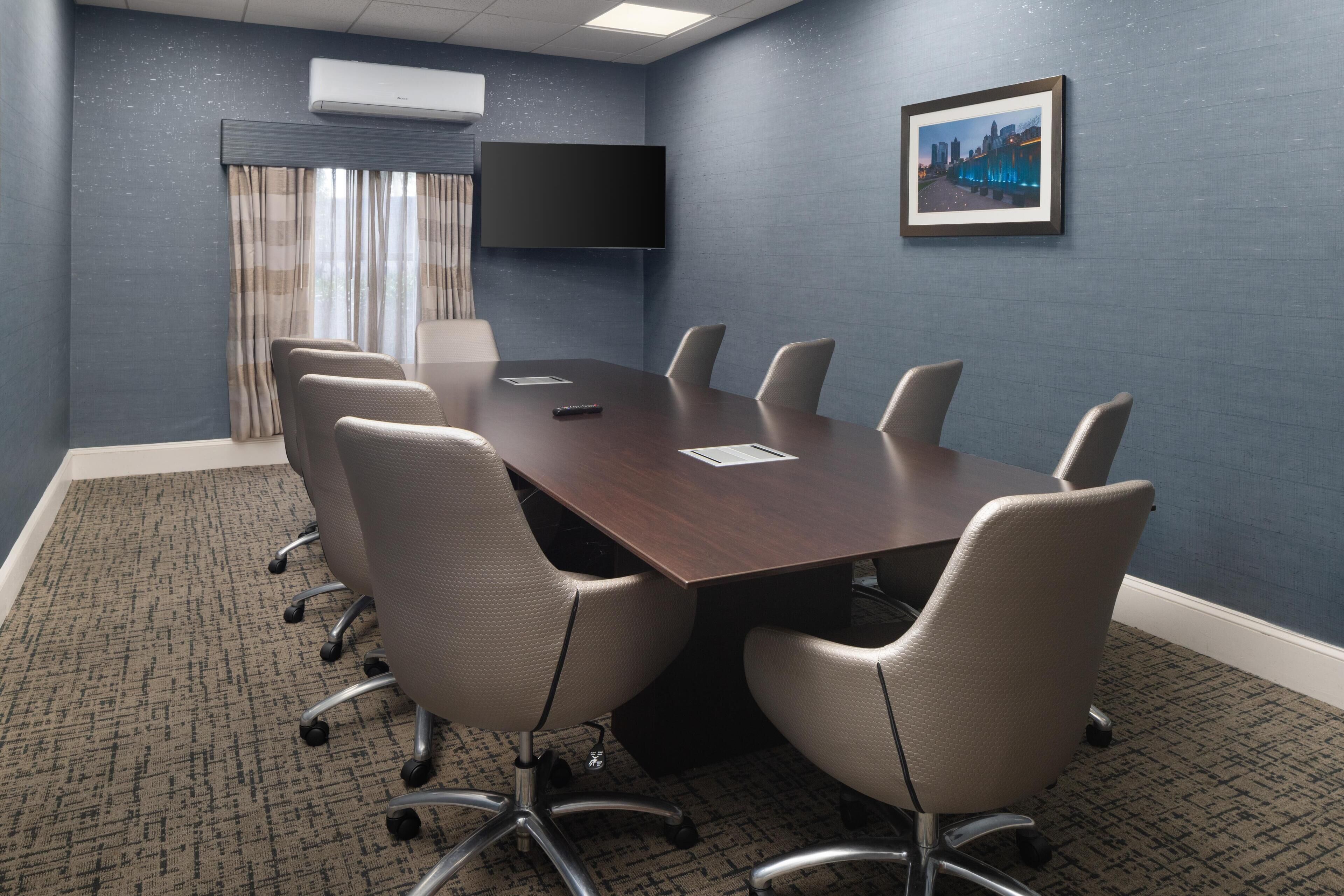 A conference room with a dark wooden table, six chairs, a flat-screen TV, and a framed cityscape picture on the wall.