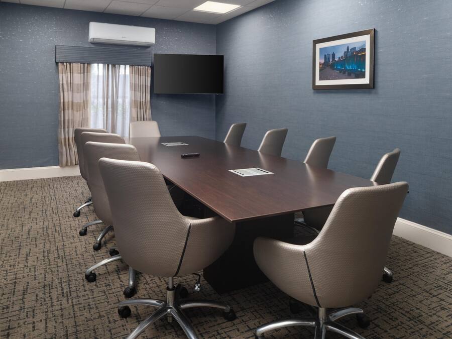 A conference room with a dark wooden table, six chairs, a flat-screen TV, and a framed cityscape picture on the wall.