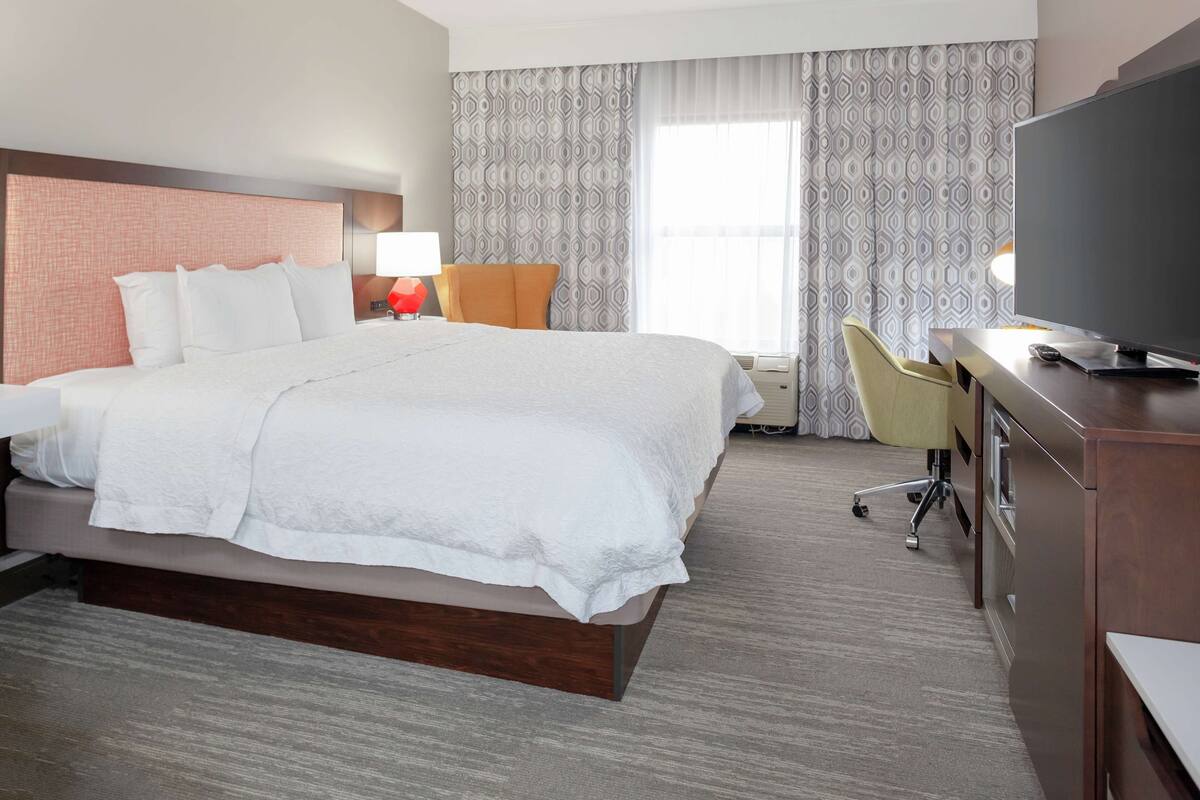A hotel room with a large bed, a desk with a television, a chair, and a window with patterned curtains.