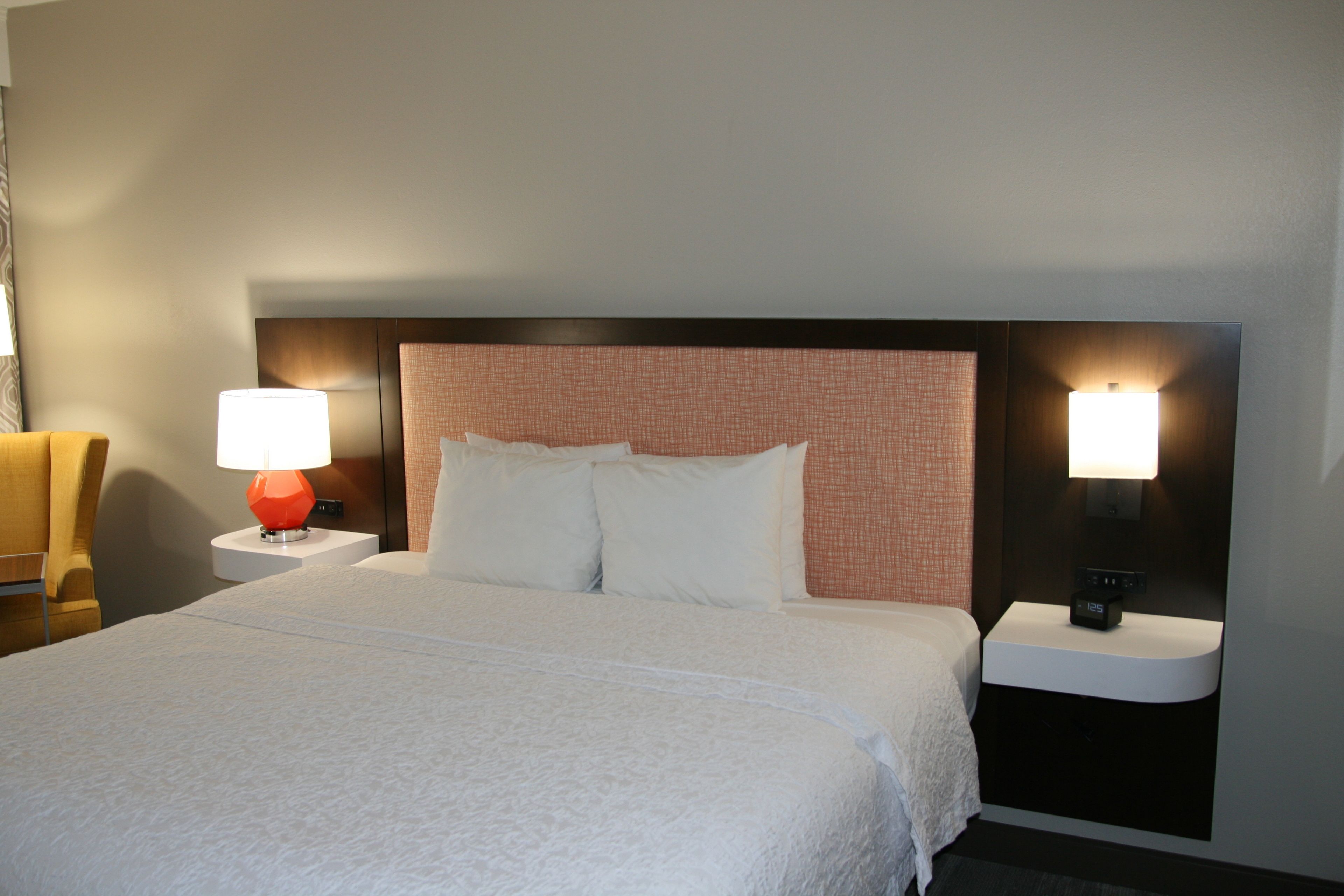 A hotel room with a large bed, two bedside tables, a red lamp, and a nightstand with a clock.