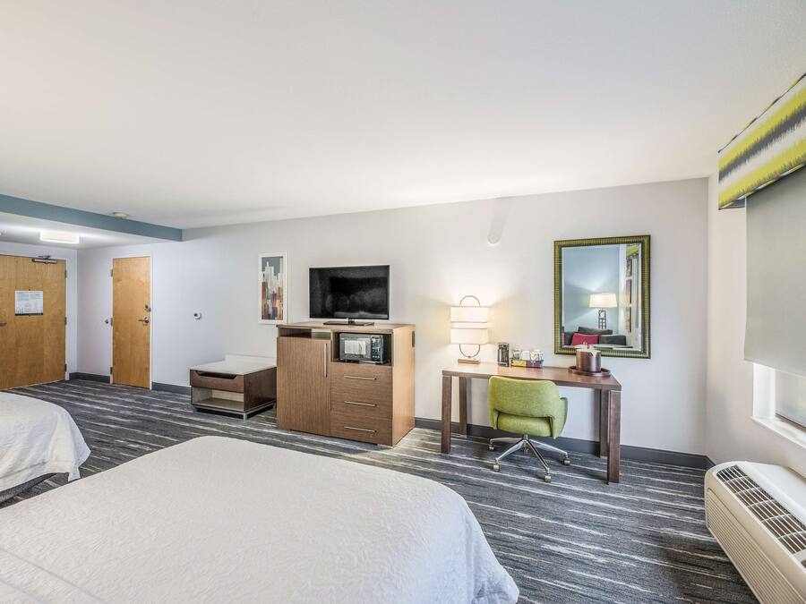 Una habitación de hotel con dos camas, un escritorio con computadora, un televisor, un espejo y una ventana.