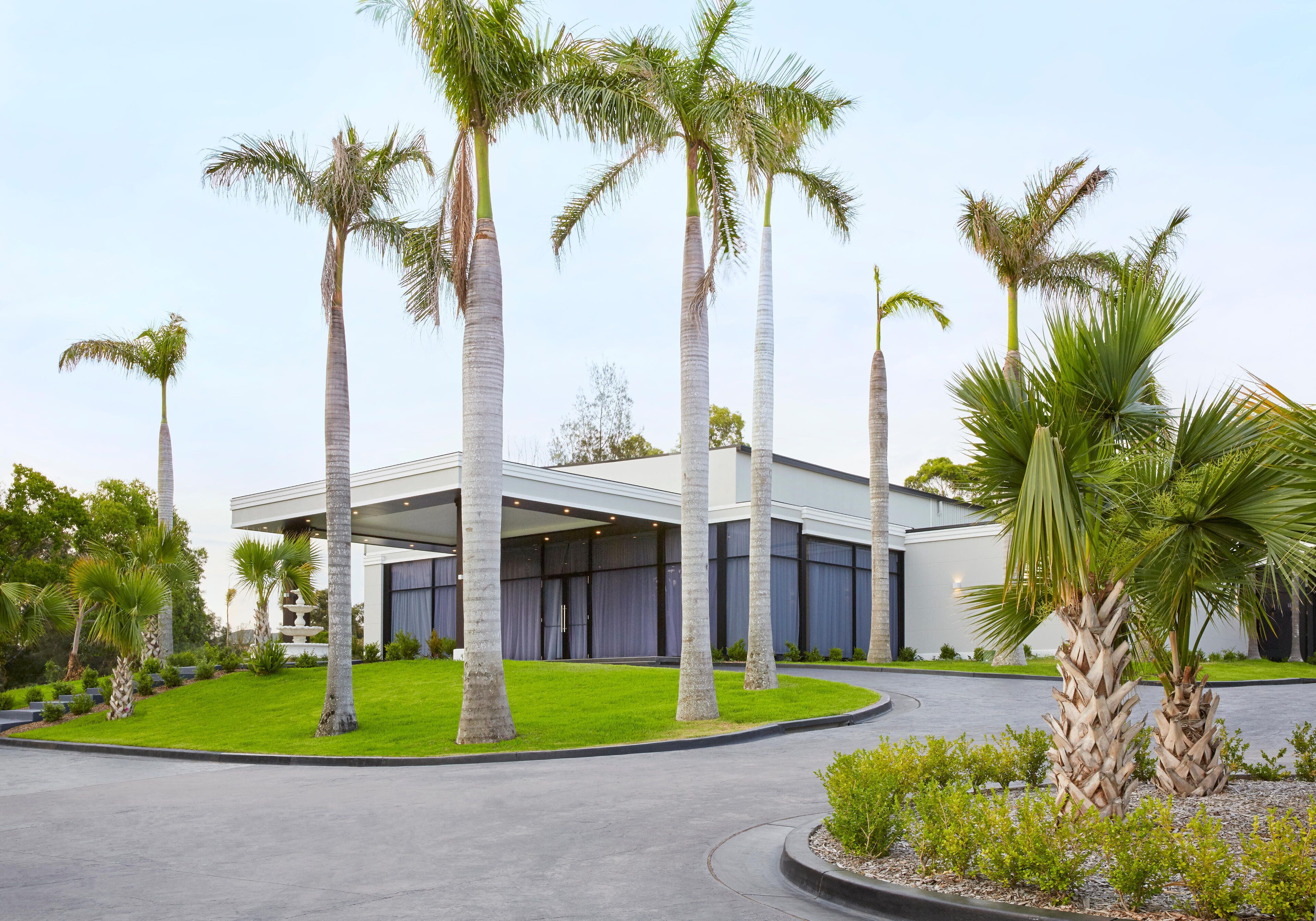 A modern building with a covered outdoor area, surrounded by palm trees and a well-maintained lawn.
