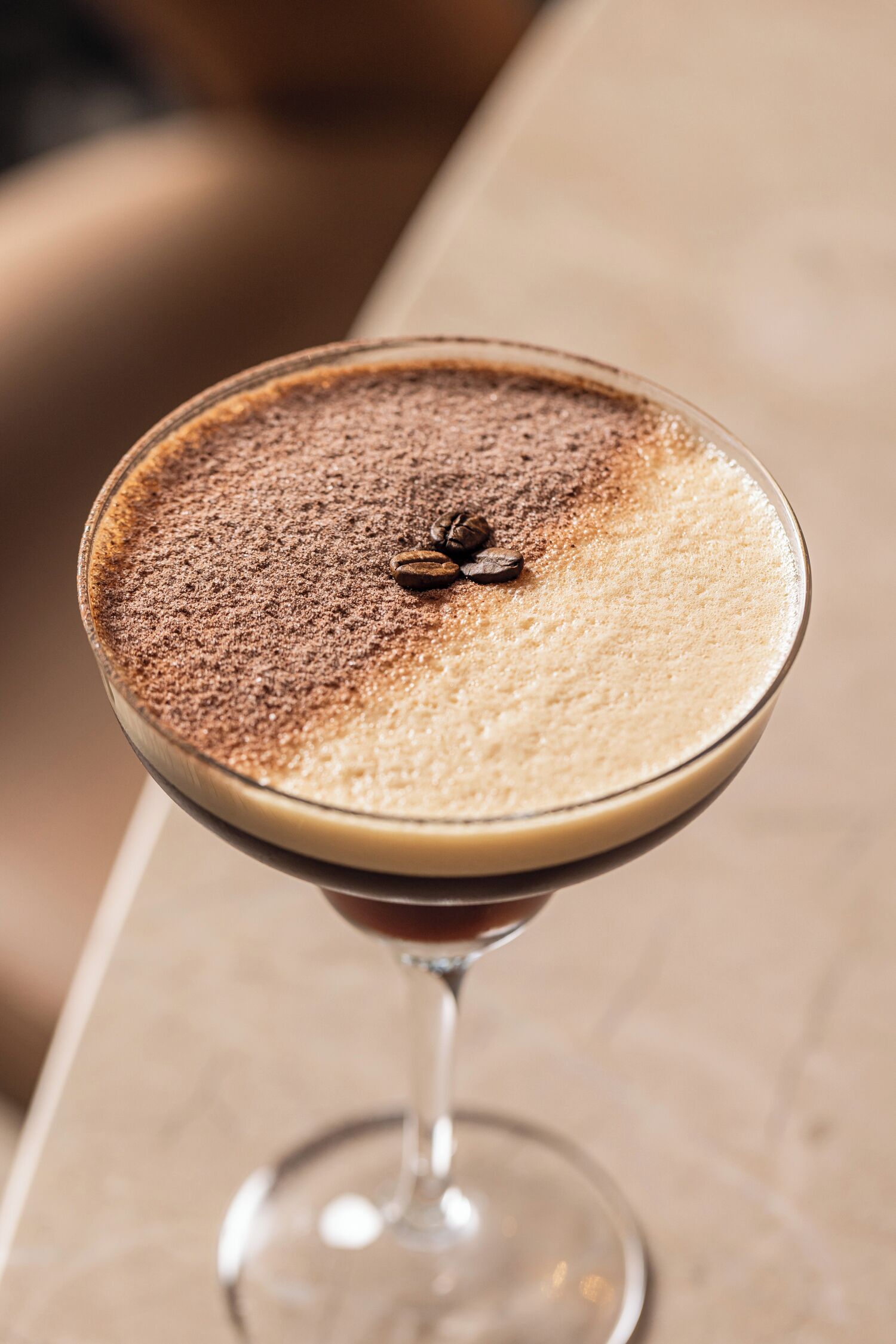 Una copa de martini con una espuma en la parte superior, un poco de cacao en polvo y un solo grano de café en el borde.