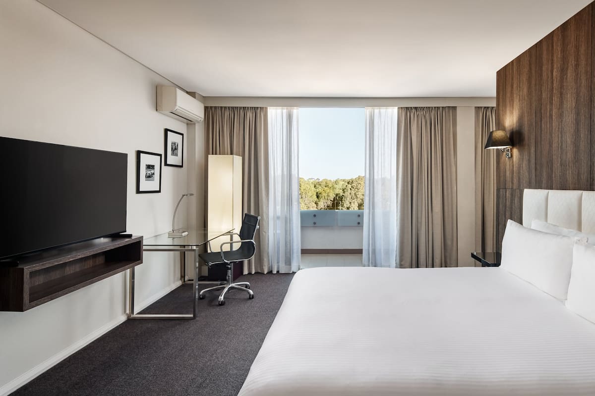 A hotel room with a large bed, a flat-screen TV, a desk with a chair, a window with curtains, and a view of trees.