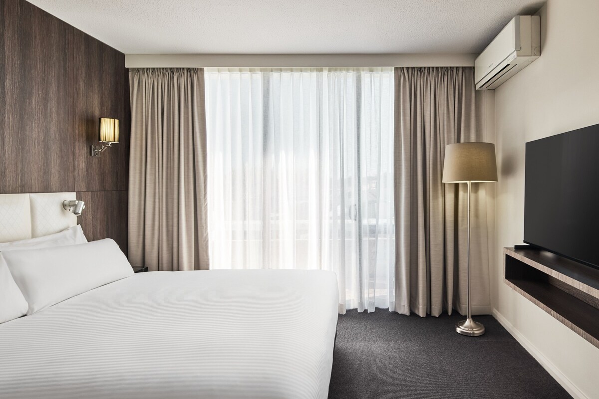A hotel room with a large bed, a flat-screen TV, a lamp, and a window with curtains.