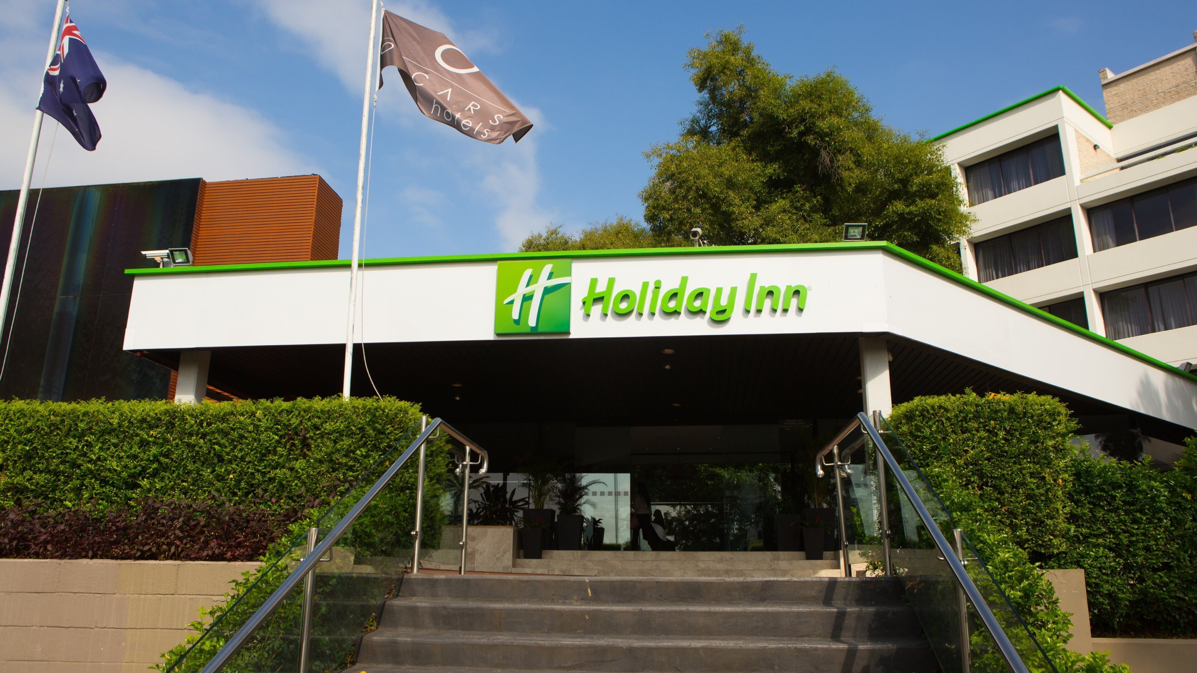 A Holiday Inn hotel entrance with a green and white facade, a set of stairs leading up to the entrance, and a flagpole with a flag.