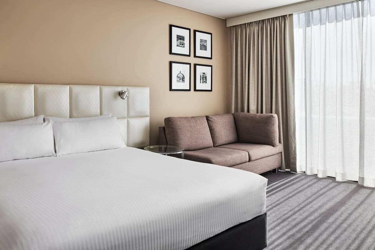 A hotel room with a large bed, a sofa, and a small table. There are framed pictures on the wall and a window with curtains.