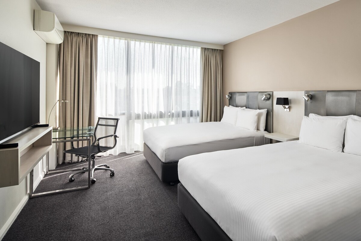 A hotel room with two beds, a desk with a chair, a television, and a large window with curtains.