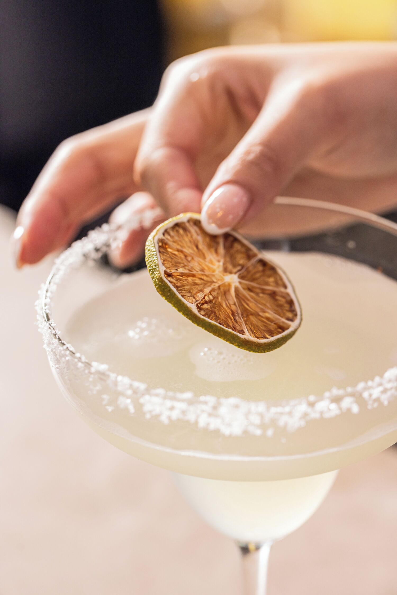 A hand holding a dried lime slice over a margarita glass.