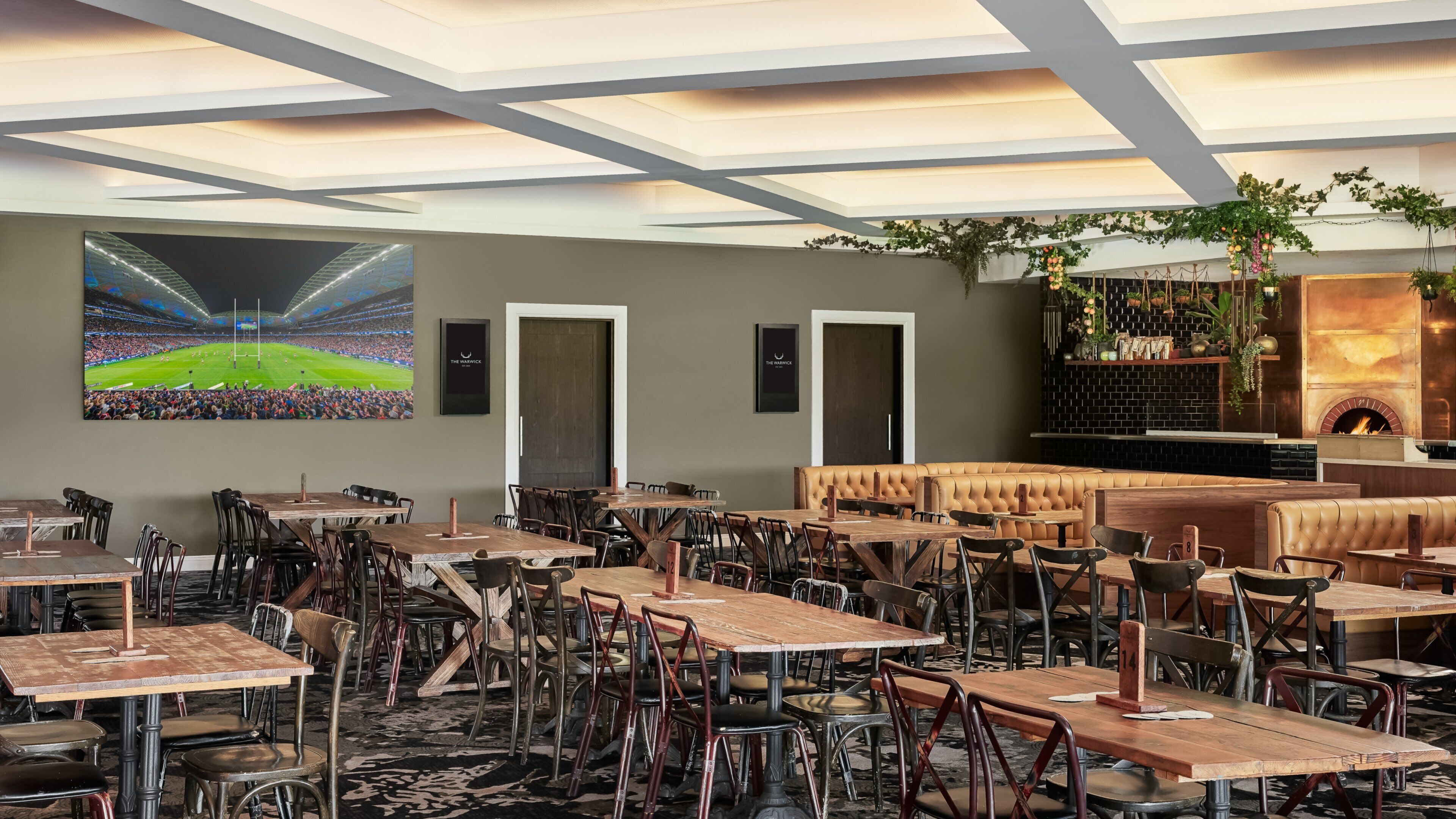 A restaurant with multiple tables and chairs, a large screen displaying a sports event, and a fireplace with plants.
