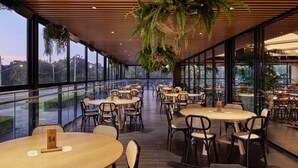 A spacious indoor dining area with multiple tables and chairs, large windows offering views of the outside, and hanging plants. - Holiday Inn Warwick Farm by IHG (Cabramatta)