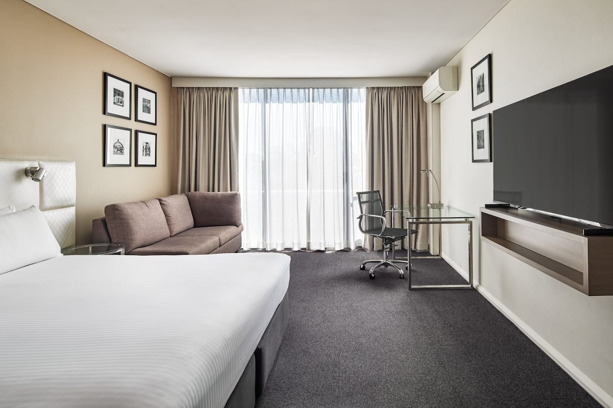 A hotel room with a large bed, a sofa, a desk with a chair, a TV, and a window with curtains.