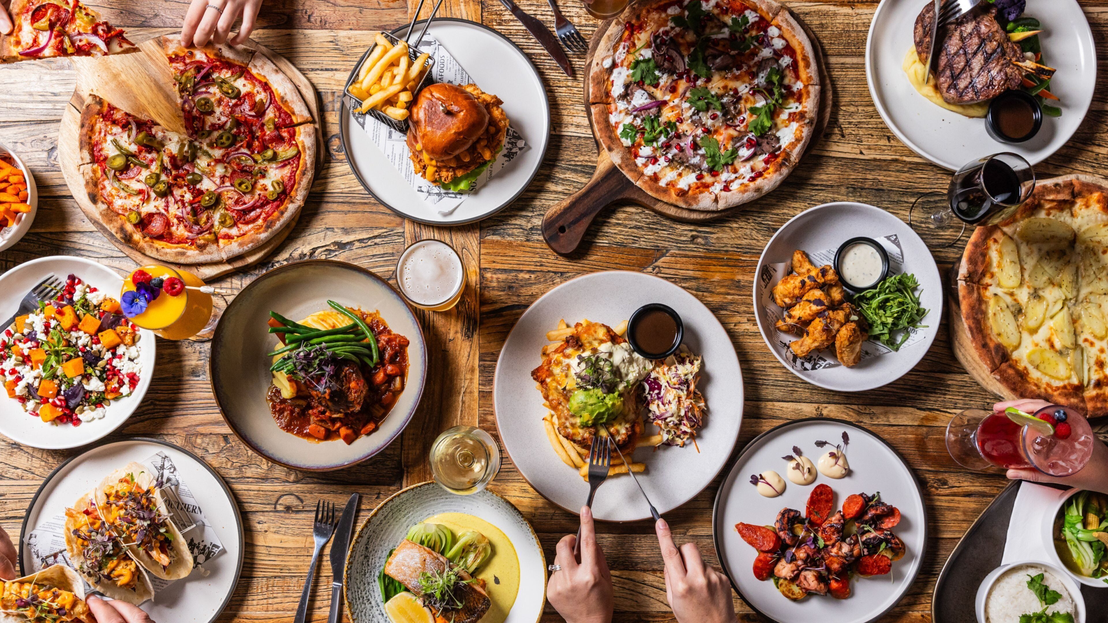 A variety of dishes on a wooden table, including pizzas, salads, and grilled meats.