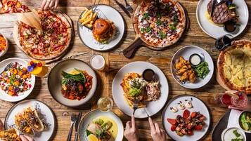 A variety of dishes on a wooden table, including pizzas, salads, and grilled meats.