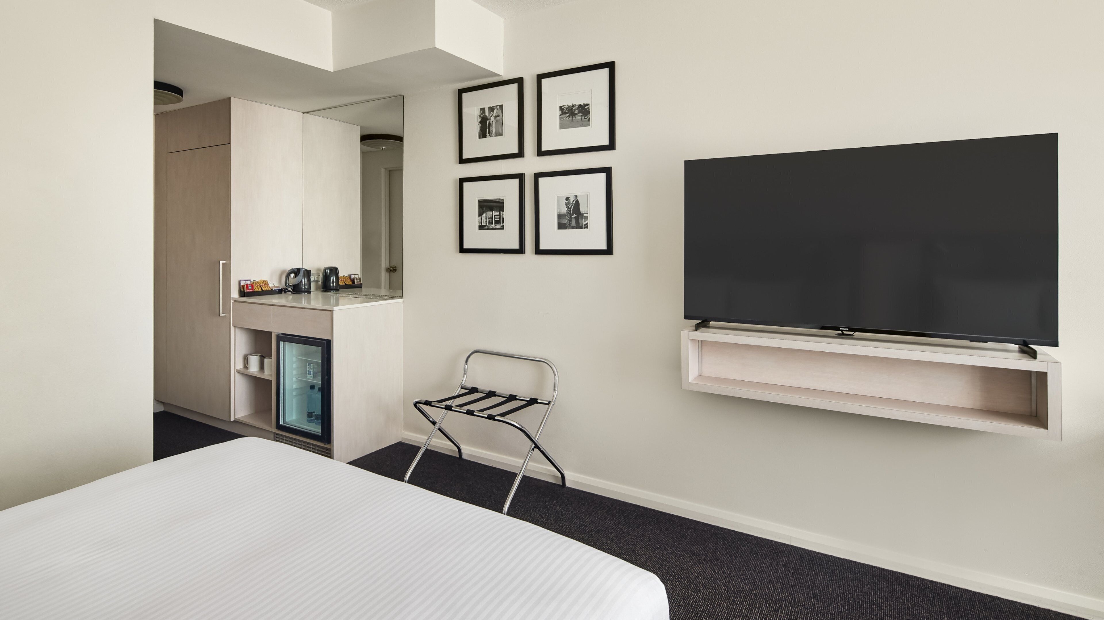 A hotel room with a bed, a TV mounted on the wall, a small table with a kettle and cups, a chair, and a refrigerator.
