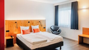 Quarto de hotel moderno com cabeceira de madeira, uma cama grande com lençóis brancos, uma cadeira preta e um banco de madeira.