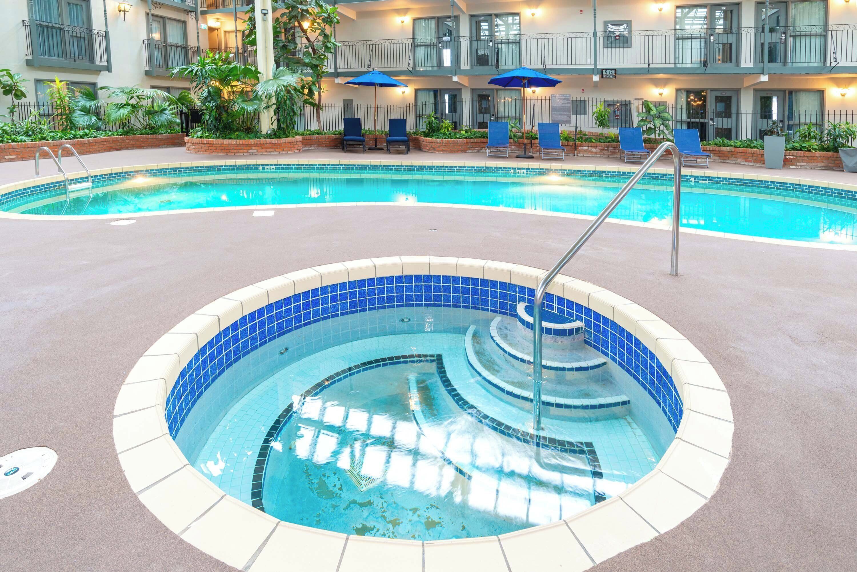 A swimming pool with a hot tub, surrounded by lounge chairs and umbrellas, with a building in the background.