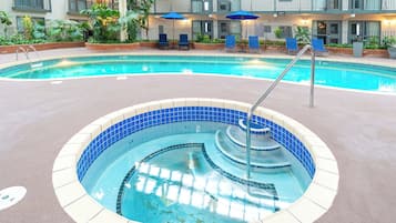 En swimmingpool med ett bubbelbad, omgiven av solstolar och parasoller, med en byggnad i bakgrunden.
