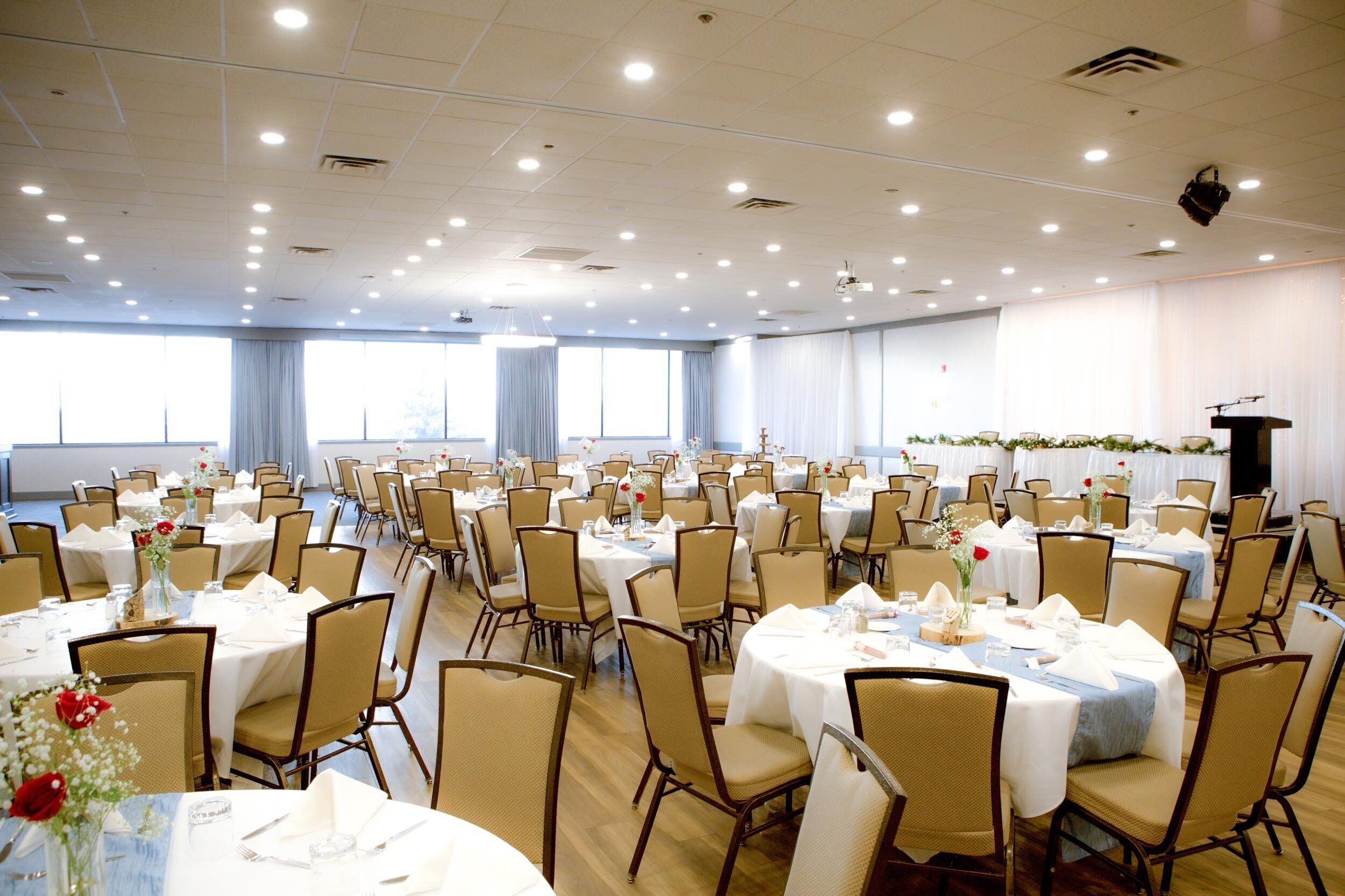 A large banquet hall with multiple round tables, each set with white tablecloths, chairs, and floral centerpieces.