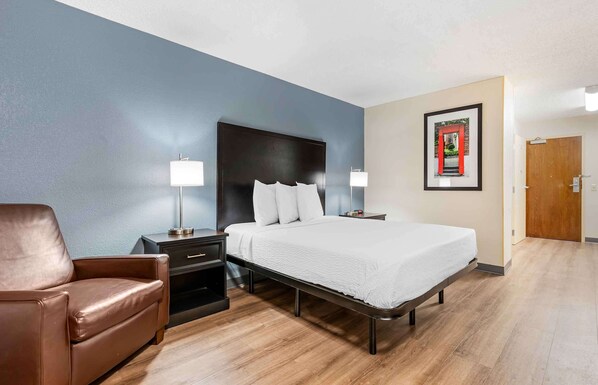 Una habitación de hotel con una cama, un sillón de cuero marrón, una mesita de noche con lámpara y un cuadro en la pared.