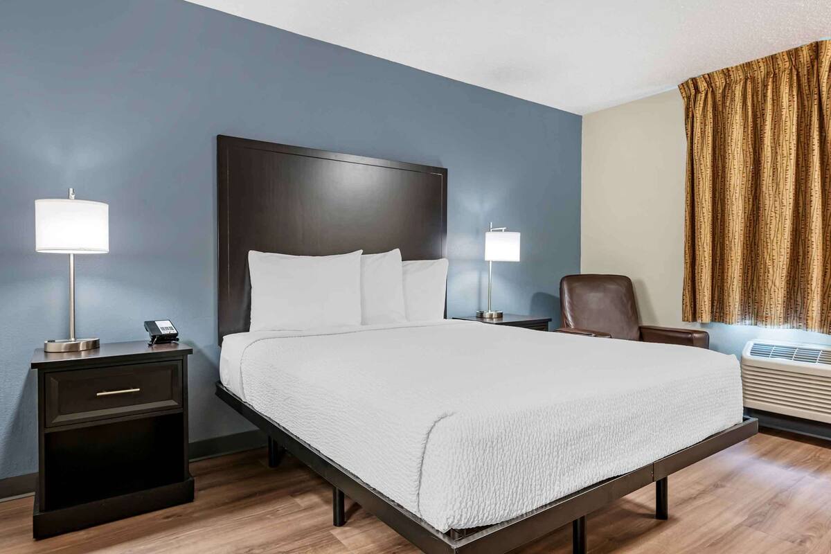 A hotel room with a large bed, two bedside tables with lamps, a chair, and an air conditioning unit.