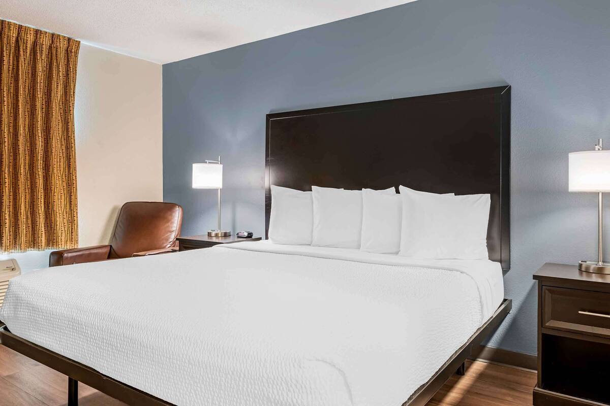 A hotel room with a large bed, a chair, two bedside tables with lamps, and a window with curtains.