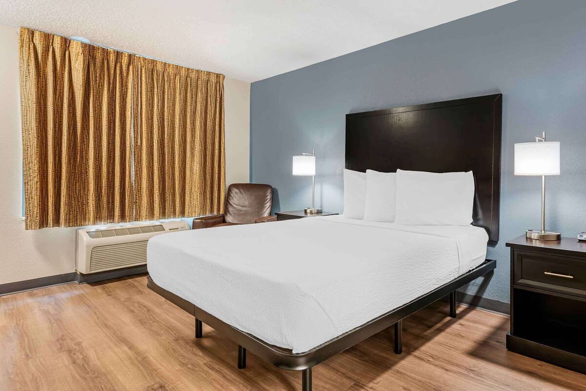 A hotel room with a bed, two bedside lamps, a chair, a nightstand, and a wall-mounted heater.