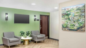 Un área de recepción con dos sillones grises, una pequeña mesa redonda con base dorada y dos plantas en macetas. Un cuadro grande que representa un paisaje adorna la pared.