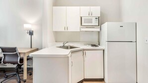 Una cocina compacta con armarios blancos, microondas, refrigerador, pileta y una silla.