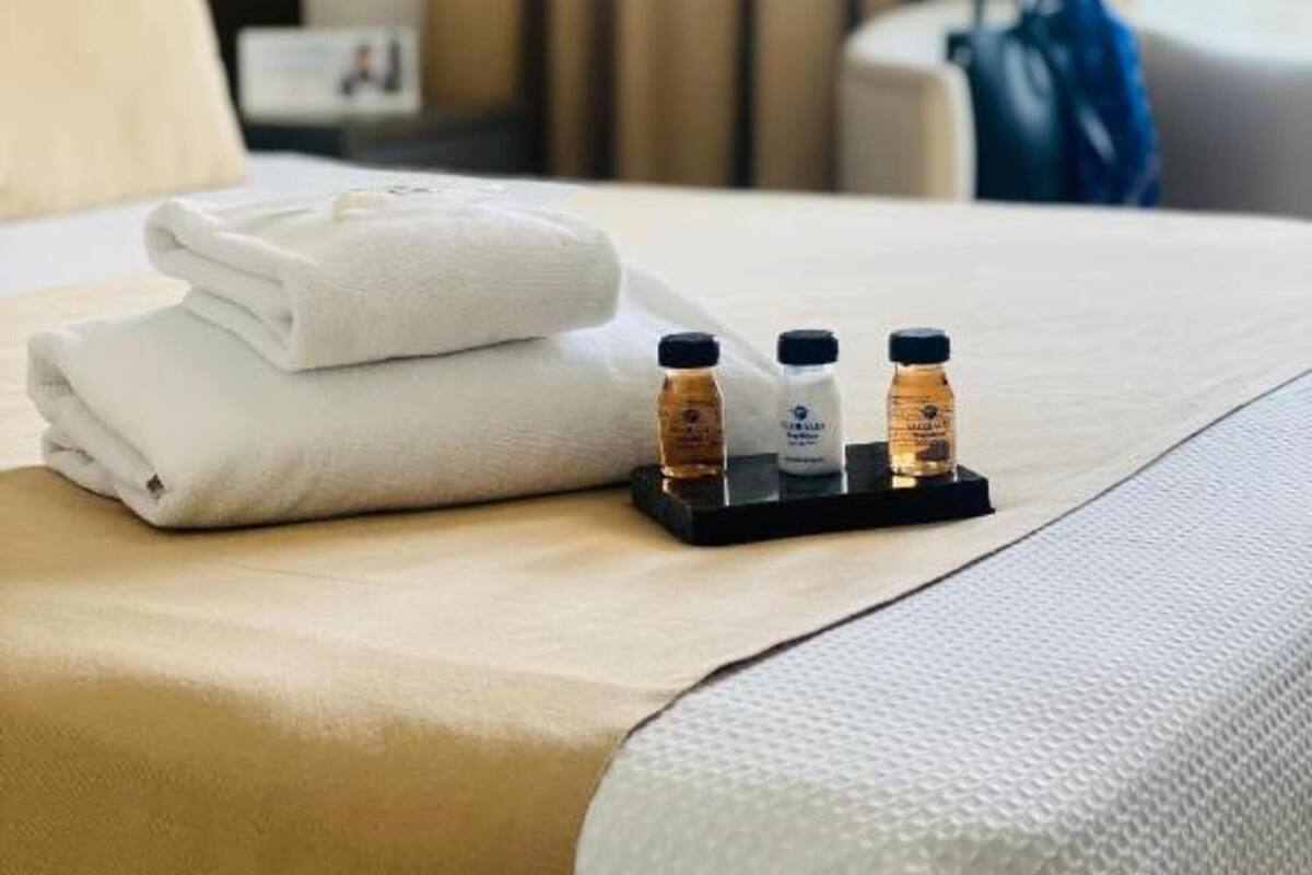 A neatly made bed with folded towels and two small bottles on a tray, a lamp, and a blue bag on a chair.