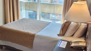 Ein Hotelzimmer mit zwei Betten, einem Nachttisch mit Lampe und Blick auf die Stadt durch große Fenster.