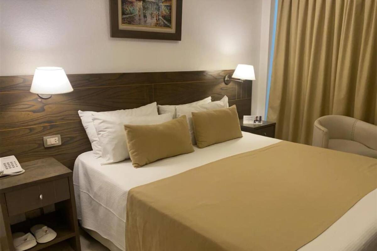 A hotel room with a bed, two bedside lamps, a nightstand with a phone, and a pair of slippers.
