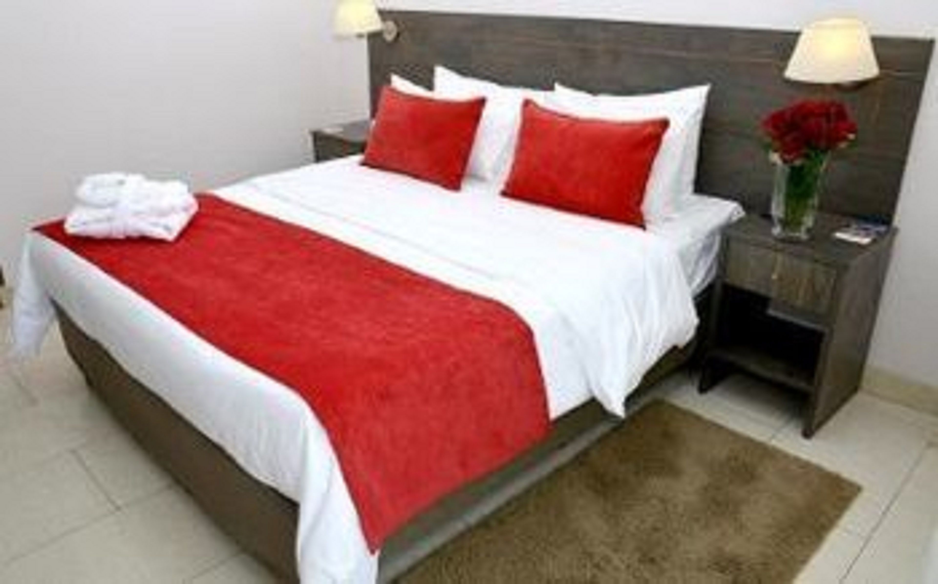 Une chambre d’hôtel avec un lit, une table de chevet, une lampe et un vase contenant une fleur rouge.
