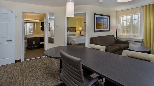 A hotel room with a dining table, two chairs, a sofa, a small round table, a lamp, a bed, a bathroom, and a window with curtains.