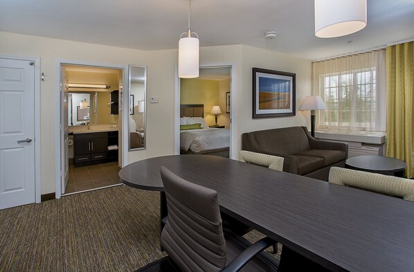 A hotel room with a dining table, two chairs, a sofa, a small round table, a lamp, a bed, a bathroom, and a window with curtains. - Candlewood Suites Louisville Airport by IHG (Louisville)