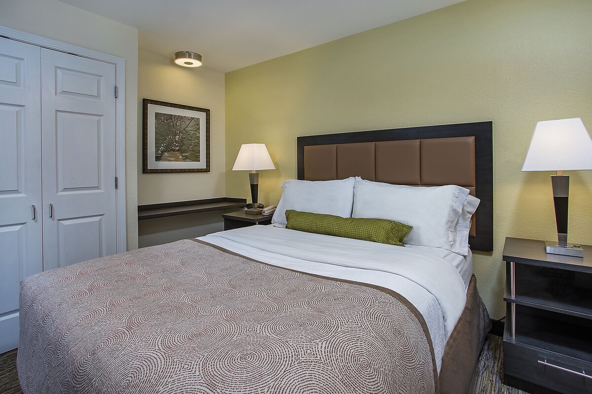 Una habitación de hotel con una cama bien hecha, una mesita de noche con lámpara, un cuadro en la pared y una puerta a la izquierda.