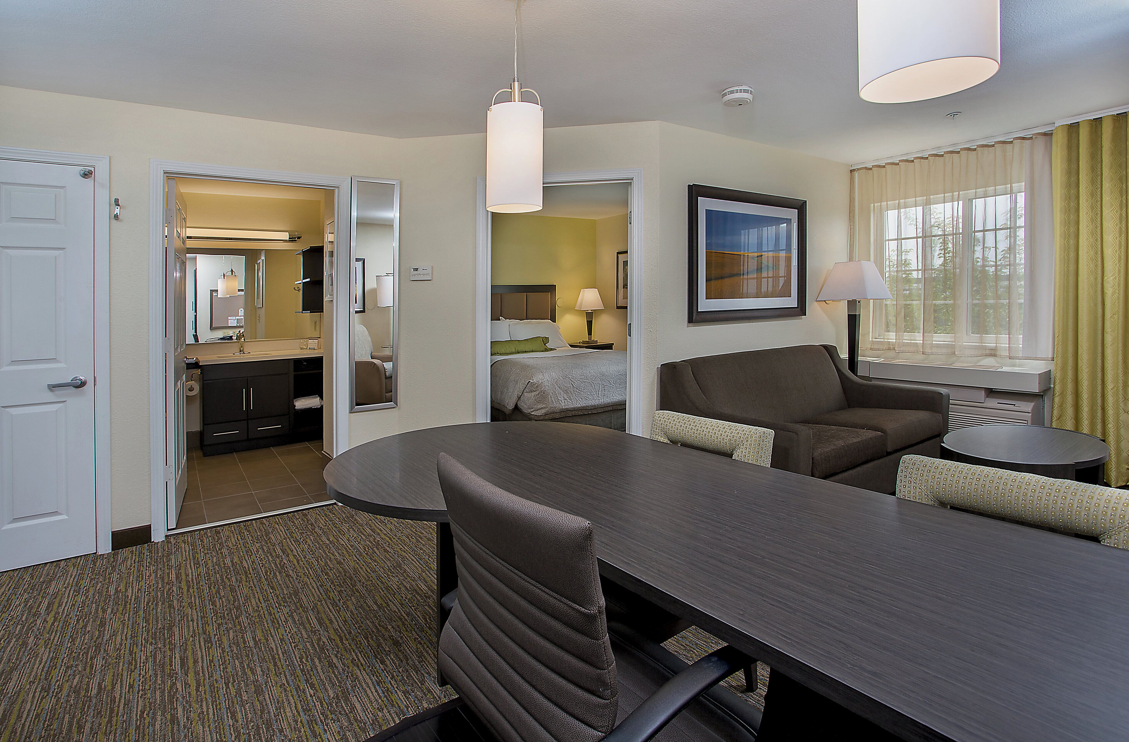 A hotel room with a dining table, chairs, a sofa, a bed, a bathroom, and a window with curtains.