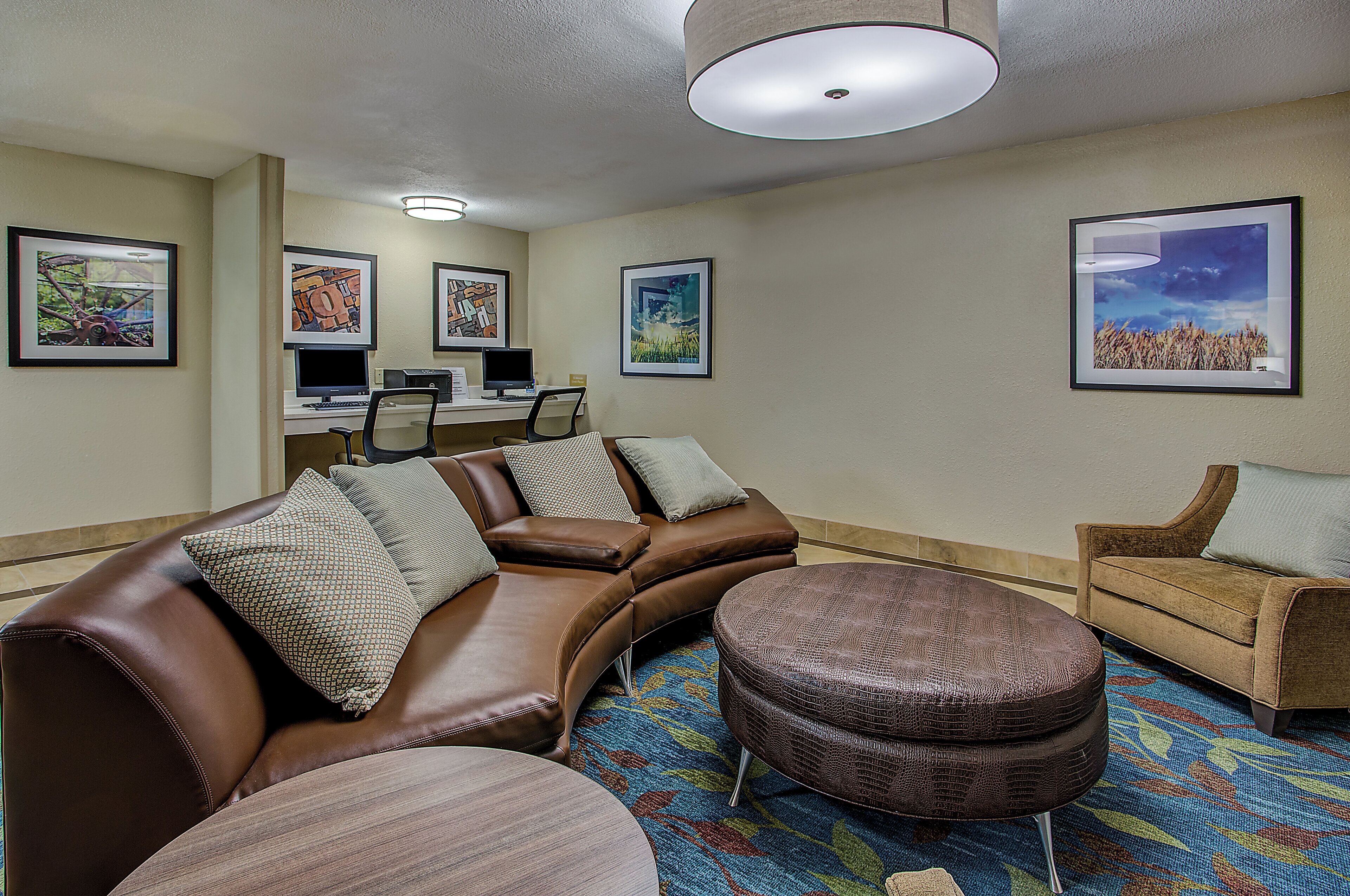 A hotel room with a brown leather sofa, a round ottoman, a small armchair, a desk with a computer, and framed artwork on the walls.